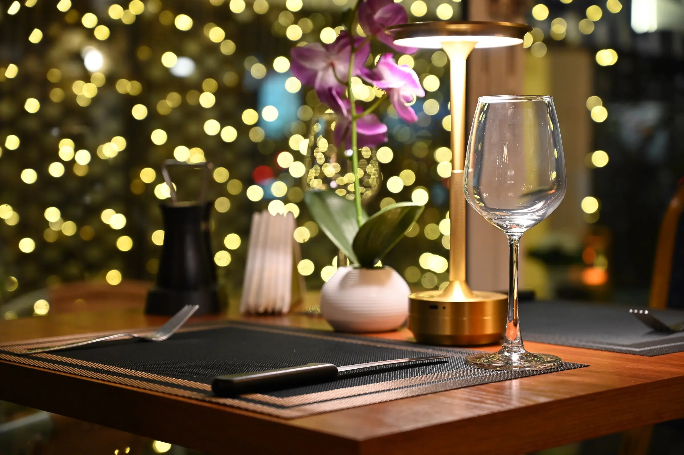 Mesa para dos con lámpara dorada, copa de vino y luces al fondo